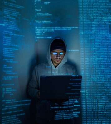 Confident young man in hooded shirt using computer while standing against dark background